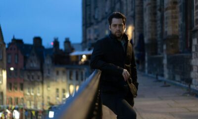 Richard Rankin as John Rebus, wearing a leather jacket and leaning on a wall