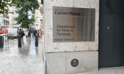 A silver sign on a wall says Welcome to Caxton House, Department for Work and Pensions, Visitors Entrance, with people walking towards the camera on the pavement to the left