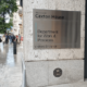 A silver sign on a wall says Welcome to Caxton House, Department for Work and Pensions, Visitors Entrance, with people walking towards the camera on the pavement to the left