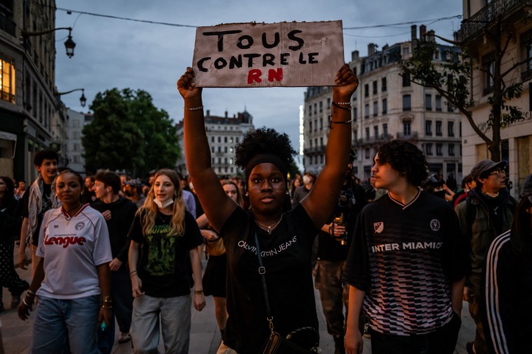 A protester in Lyon holds up a placard reading 'Everyone against the RN'. There are people walking behind her.