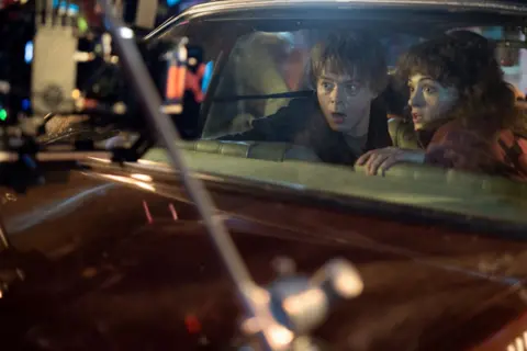 Netflix A behind-the-scenes shot of a young male and female actor in the back seat of a vintage car. They're looking out of the back window, with shocked expressions on their faces. In the foreground a camera rig capturing the pair's faces is just about visible.