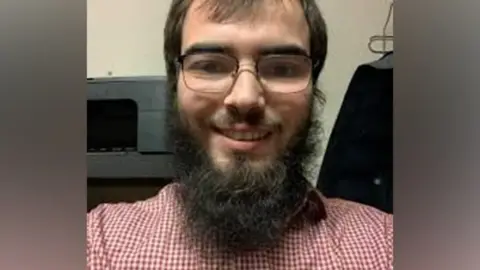 Met Police  Khaled Hussein has brown hair, a large beard and glasses and a red and white shirt - the picture appears to be a selfie and he is smiling into the camera 