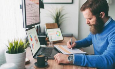 Bearded man writing on notepad in front of computer