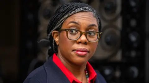 BBC Kemi Badenoch wearing glasses and a red blouse looks at the media via the corner of her eye in Downing Street