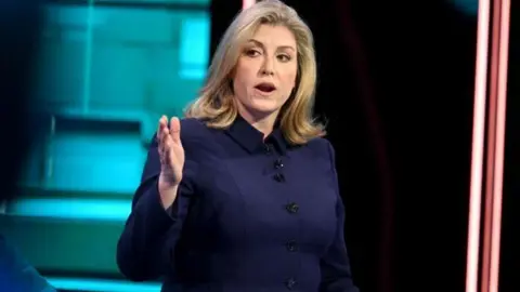 ITV/EPA Penny Mordaunt in a navy blue suit, looking to her left with arm and hand extended. She has shoulder-length blonde hair