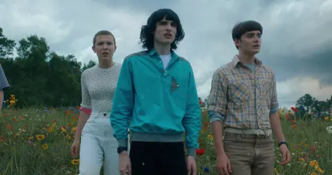 Netflix Three young people, two males and a female with a shaved head, stand in a field with long grass and various colours. They're looking out at something off-camera in front of them, with expressions of concern and worry.