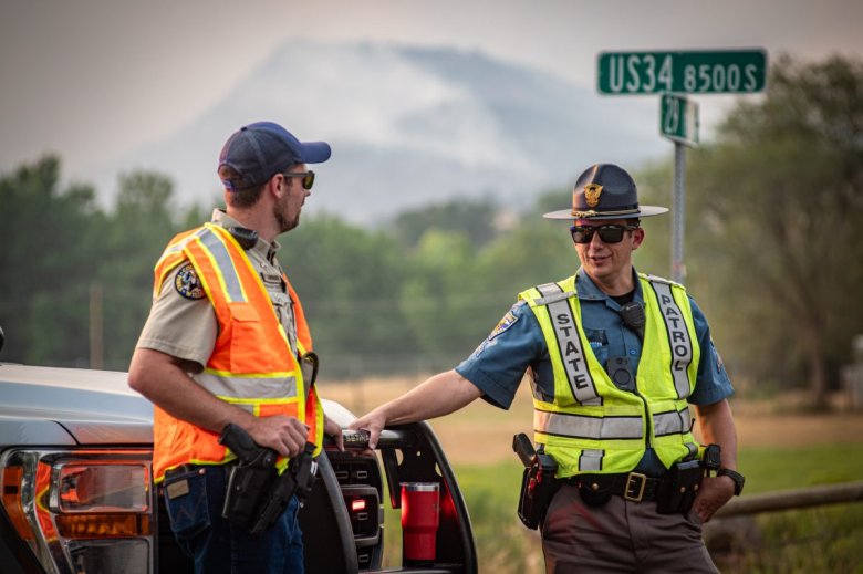 Two officers wearing reflective vests converse near a patrol vehicle, with a road sign marked 