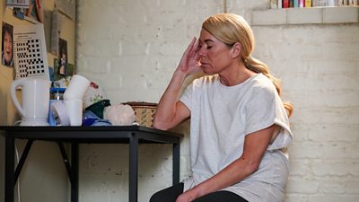 Sharon sitting a table in a prison cell with her hand to her head and eyes closed