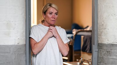 Character Sharon in the doorway of a prison cell clutching her hands to her chest and looking sad/worried. She's got her hair in a low ponytail and no makeup 