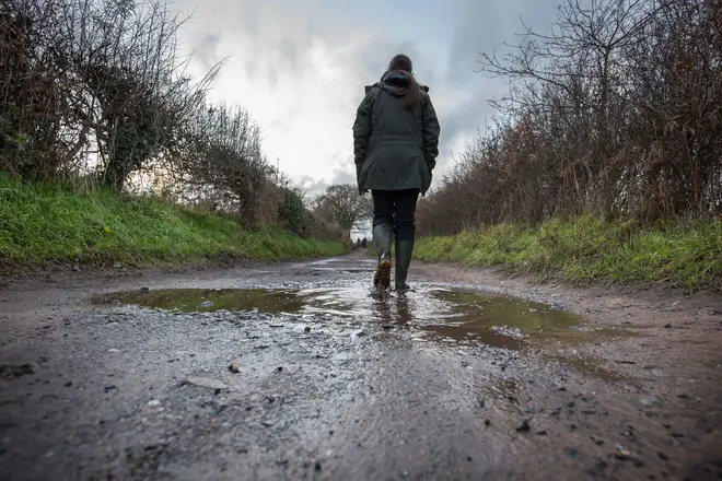 Low angle, rear view of an isolated woman wearing green wellies & coat, walking down a wet, muddy, UK country lane on a December afternoon, 2020.