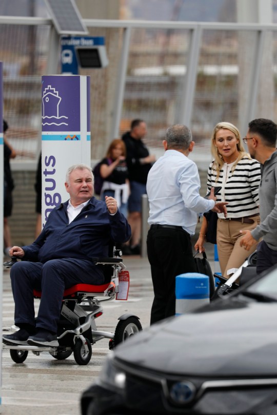 Eamonn Holmes wearing a navy blue jacket and trousers and using a wheelchair at Barcelona airport 