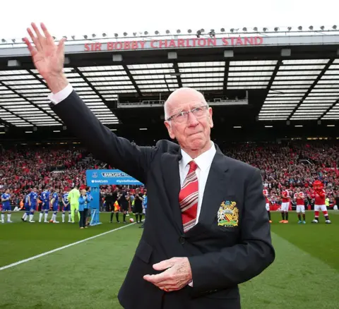 Getty Images Sir Bobby Charlton, wearing a black blazer, white shirt and red striped tie, stands on the pitch at Old Trafford waving to crowds. He is attending the unveiling of a stand renamed in his honour, which can be seen packed with fans in the background