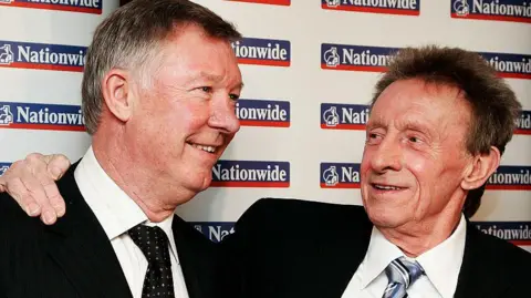 Getty Images Denis Law, wearing a black suit, white shirt, and blue and grey striped tie, stands with his arm around Sir Alex Ferguson, wearing a black suit, white striped shirt and black tie