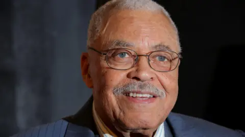 Getty Images James Earl Jones wearing round spectacles looks towards the direction of a camera and smiling. The actor has a grey moustache and short grey hair. 