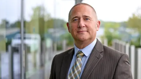 Martismedia Detective Stuart Brooks, from Avon and Somerset Police, looks at the camera. He is wearing a grey pin-striped suit, light blue shirt and a cross-patterned tie.