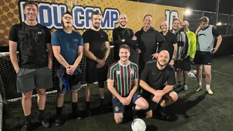 A group of male footballers line up for the camera in a group formation at Goals Bristol South in Brislington. Two of them are kneeling at the front with a football at their feet and many of the players are smiling. The picture is taken at night and there are floodlights on in the background