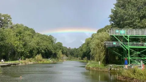 Alison Kay Henleaze Lake is pictured looking down its length from one end, with trees and grassy areas on each side. Some people are swimming in the water and a young woman is standing on the lowest of its three diving boards. There is a rainbow overhead