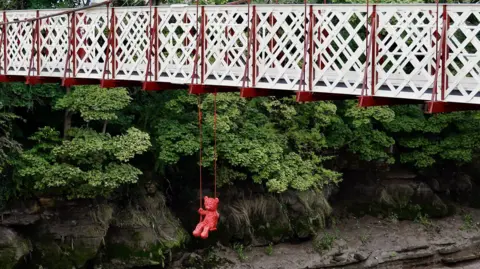 James Colomina A red teddy bear street art creation hangs below Gaol Ferry Bridge in Bristol. The ropes holding the swing are also red, and in the background the muddy banks of the Avon and green trees and bushes on the banks are visible.