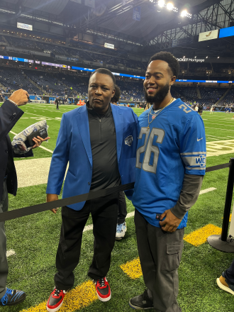 Barry Sanders and Brian Cross stand together on the field inside Ford Field in Detroit.