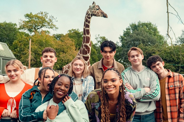 Rhea Norwood, Toby Donovan, Leila Khan, Corinna Brown, Kizzy Edgell, Will Gao, Yasmin Finney, Kit Connor, Joe Locke standing in the middle of a zoo and in front of a giraffe, smiling.