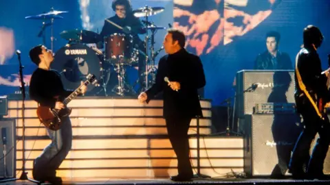 Getty Images Tom JONES on stage with STEREOPHONICS, Kelly and Sir Tom are up front together look towards each other as they sing and Kelly plays guitar. You can also see the drummer keyboardist and the bas player is looking away.