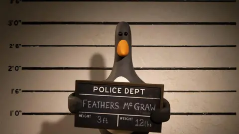 Aardman Animations Ltd / BBC Feathers McGraw, a stop-motion animated penguin, is seen holding up a police department sign while having a mugshot taken