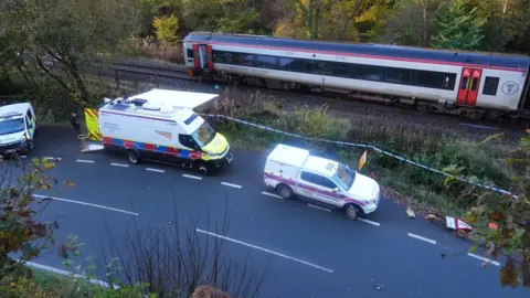 Police van, Network Rail van and Network Rail response unit jeep on the side of the road alongside the track, with the crashed train stationary on the tracks in daylight. The doors of the end carriage are open and there is police tape in front of the tracks.