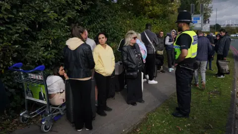 PA Media People stood on a pavement, one is sat on a luggage trolley. A police officer is stood nearby the group.