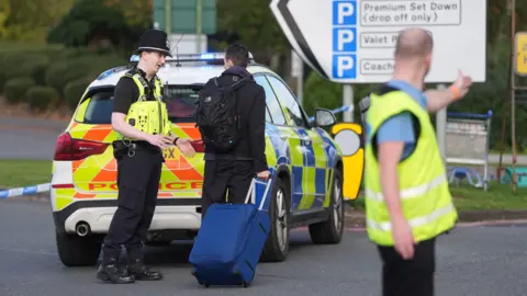 PA Media A man wheeling a suitcase talks to a police officer who is stood at the back of a police car, while another man in a hi-vis tabard is seen with is arm outstretched and appears to be giving directions.