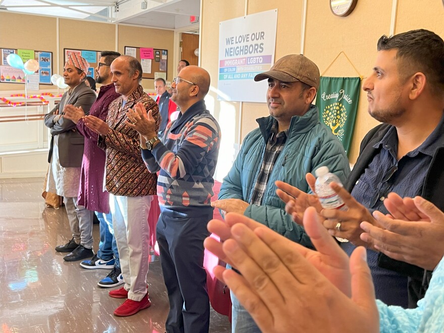 A row of men stand at the back of a room clapping to music.