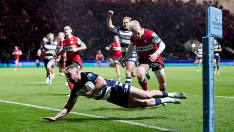 PA Media Bristol's Richard Lane dives over the line to score a try as Bristol Bears host Gloucester at Ashton Gate on Friday night. In the background one of his team-mates begins celebrating while two Gloucester players look on 