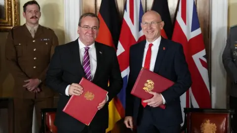 PA Media Healey and Pistorius smile, standing in front of the Union Jack and German flags, holding red binders 