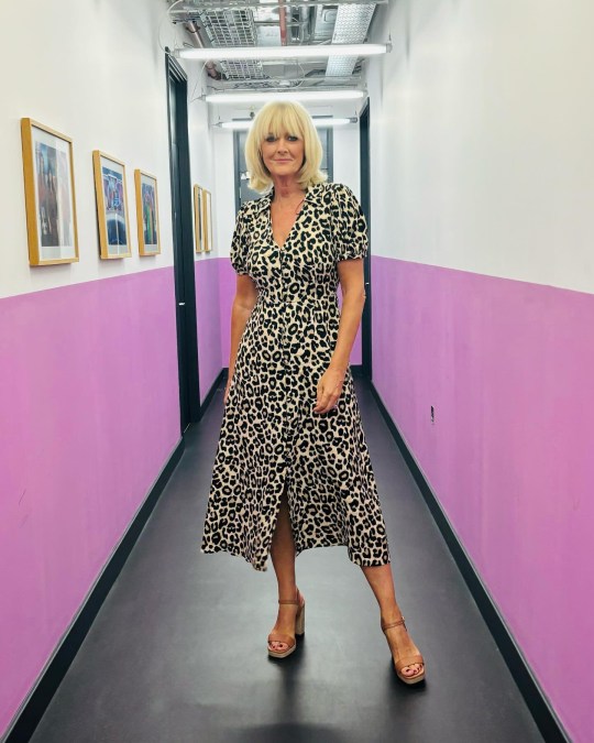 Jane Moore poses in a white and purple hallway, wearing a leopard print dress and brown wedge heels