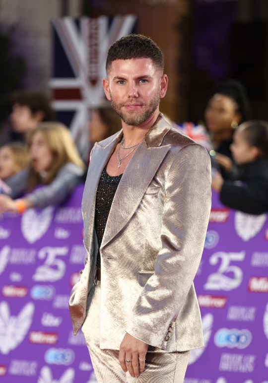 Dean McCullough poses on the red carpet wearing a gold suit and black sparkly top