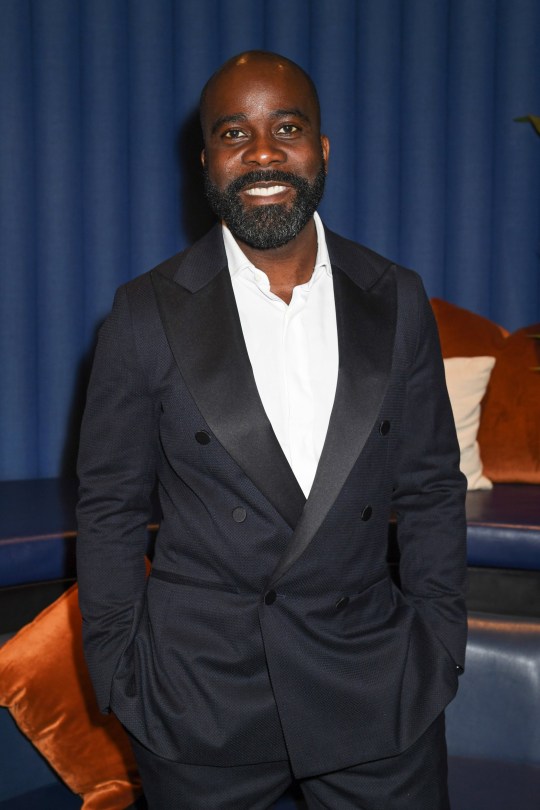 Melvin Odoom smiles as he poses in a black suit jacket and white shirt