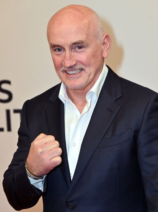 Barry McGuigan poses with his hand in a fist, while wearing a black suit jacket and white shirt