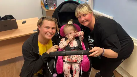 Tom Walker/BBC Three-year-old Robyn, with pink glasses and a pink flowery outfit, sits in a pram next to her smiling brother Josh, who is on the left and has blonde hair pushed back with a hairband, and her mother Stephanie Perry, who has a black t-shirt and blonde hair in a fringe