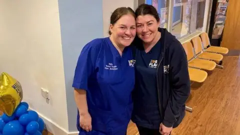 Tom Walker/BBC Katy Jones and Laura Knight, in blue scrubs, smile and pose for the camera with their faces touching 