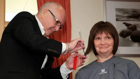 Getty Images Trevor Sorbie styling the wig of a cancer patient. He is wearing a black jacket and white shirt and has a bald head and glasses. He is using scissors to style a brown wig on the head of a woman who is wearing a grey Trevor Sorbie hairdressing cape. He is also holding some red scissors and has a chunky bracelet on his wrist. There is a red curtain and a black and white framed picture in the background.