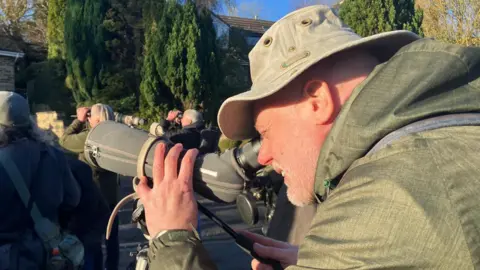 BBC / Elizabeth Baines Paul, dressed in green, peers through a long lens to see the Scarlet Tanager.