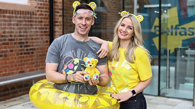 Connor Phillips and Holly Hamilton smile to camera wearing branded clothing for BBC Children in Need. Connor wears goggles and an inflatable rubber ring in reference to the ‘mammoth challenge swim team’ challenge. 