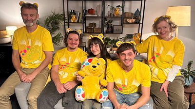 The Brockmans sit on a grey sofa wearing Children in Need t-shirts and Pudsey bear ear headbands, holding a large Pudsey bear stuffed toy