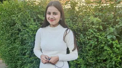 Northamptonshire Police Harshita Brella smiles at the camera as she is photographed outside. There is a large green hedge behind her. She is wearing a beige polo-necked jumper and has long dark hair. She is wearing earrings.