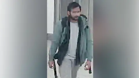 Northamptonshire Police CCTV of Pankaj Lamba wearing a green coat, white top and grey pants. He looks to have a backpack and is walking. 