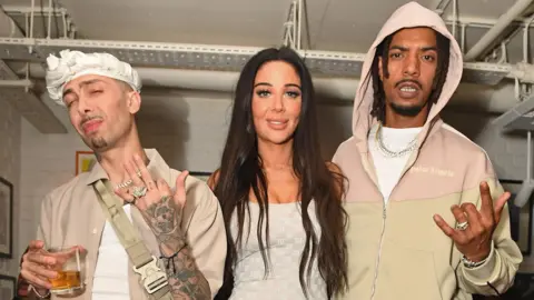 Getty Images Dappy aka Costadinos Contostavlos, Tulisa aka Tulisa Contostavlos and Fazer aka Richard Rawson of N-Dubz pictured at an event in 2023. They are all dressed in neutral colours and pictured inside a room with painted white bricks. 