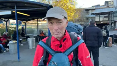 An elderly man wearing a baseball cap, red jacket and blue rucksack on his front