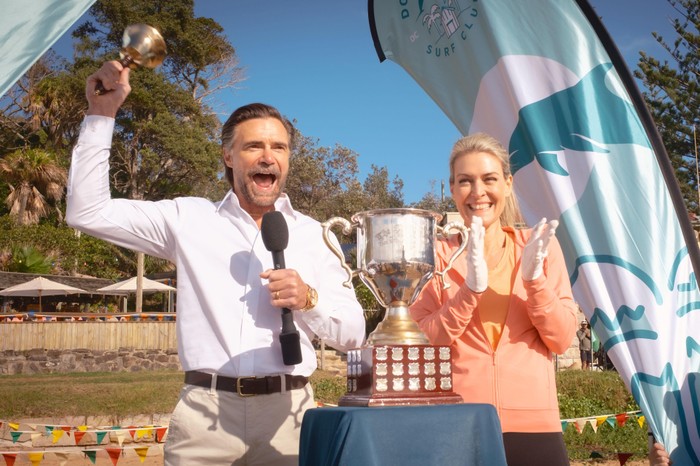 Jeremy Lindsay Taylor plays Stuart Granger and Jenni Baird plays Susan Sterling, stood next to one another on a beach. He's ringing a bell and she had a big silver cup placed in front of them