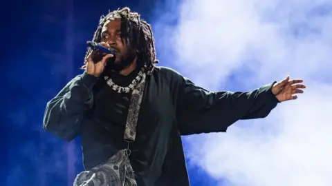 Getty Images Rapper Kendrick Lamar, wearing black clothes, a necklace, and a bag over his shoulder, holds a microphone as he performs onstage during day three of Rolling Loud Miami 2022 at Hard Rock Stadium on July 24, 2022 in Miami Gardens, Florida