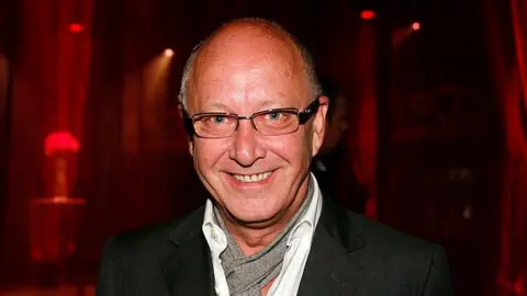 Getty Images Trevor Sorbie, who has a bald head and glasses, smiles at the camera. He is wearing a black jacket, white shirt and a speckled grey cravate. There is a dark red background.