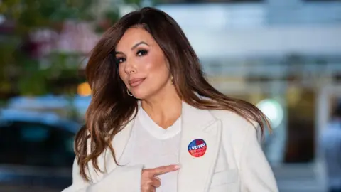 Getty Images Eva Longoria, who has long brown hair and is wearing hoop earrings, a white trench coat and a plain white T-shirt, points with her right index finger to an 'I Voted' sticker over her left lapel